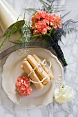 Sponge fingers tied with string and salmon-pink carnations on white plate next to bouquet of carnations on marble surface