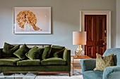 Sofa mit grünem Samtstoffbezug vor Wand mit Bild, seitlich Tischleuchte auf Beistelltisch in edlem Wohnbereich