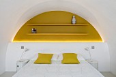 Yellow scatter cushions on double bed against half-height white wall below yellow-painted floating shelves in arched yellow niche with indirect lighting