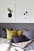 Scatter cushions arranged against grey upholstered headboard below framed pictures on white wood-clad wall