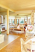 Open-plan interior with pale wooden ceiling, wicker furniture and mother and daughter in dining area in window bay
