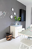 Vase of flowers on white retro chest of drawers below decorative plates on grey wall