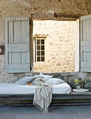 Matratze mit Kissen und Plaid unter geöffneten Fensterläden, Blick auf Natursteinmauer