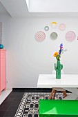 Cactus-shaped vase of flowers on white table, green bench and pastel decorative wall plates in dining room