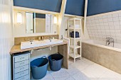 Small chest of drawers and blue baskets below simple washstand with twin sinks on wooden frame next to bathtub with concrete surround in converted attic