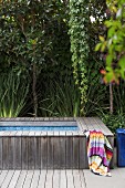 Holzverkleidete Pool-Einfassung und buntes Handtuch vor dicht gewachsenen Grünpflanzen