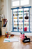Craft materials and wooden frame holding reels of string and ribbon on windowsill above desk