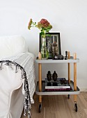 Antique picture, sedum in glass vase and ethnic ornaments on serving trolley next to white loose-covered sofa