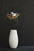 Easter arrangement of bird's nest amongst black branches in white vase against black wall