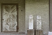 Zwei Figuren auf Holzsockeln vor Backsteinwand und Holztür