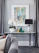 weiße Orchidee, Vasen und Fornasetti-Tischlampe auf Konsolentisch vor modernem Bild an grauer Wand