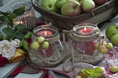 Glasses as lanterns with candles on decorative gravel, string and decorative apples