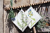Hand-made potholders with floral motifs and bunch of lavender hung from wooden rake leant against rustic board wall
