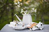 Coffee cup, books, plaited bun, and flowers in watering can on grey tablecloth