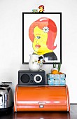 Colourful artwork above radio and fruit basket on top of old bread bin