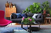 Graue Polstercouch mit blauen Kissen und pinker Sessel in Wohnbereich mit verschiedenen Grünpflanzen