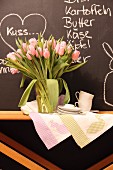 Vase of tulips, tea towels with Easter motifs and coffee service on wooden table against chalkboard with chalk messages