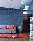 Grey, loose-covered sofa with orange scatter cushions against grey-blue stucco lustro wall with stacked books and staircase to one side
