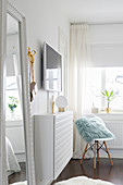 Table lamp on narrow, white, floating cabinet below wall-mounted TV and classic chair in corner below window