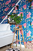 Stool next to bed under sloping ceiling covered with floral wallpaper