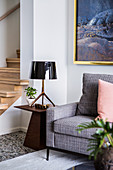 Upholstered armchair and elegant side table with lamp in front of stairs