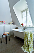 Free-standing bathtub, stool and fitted cupboards below shelf in white bathroom with dormer window