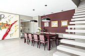 Upholstered chairs in elegant dining area with claret-red wall between huge artwork and cantilever staircase