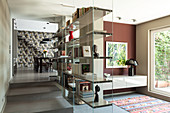 Glass partition shelves in open-plan interior with dining area and staircase in background against patterned wallpaper
