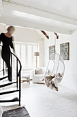 Woman walking down winding staircase into white living room with hanging chair