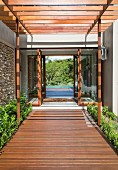 Wooden walkway below wooden pergola leading to entrance of modern house with view through to pool and trees