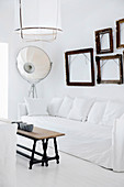 Sofa with white cover and pillows, blank picture frames above it, vintage coffee table on white plank floor