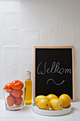 Zitrusfrüchte, Ölflasche und Willkommen-Tafel vor weißer Fliesenwand in der Küche