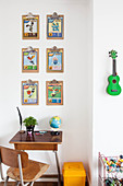 Vintage desk and chair below picture cards on clipboards and ukulele on wall