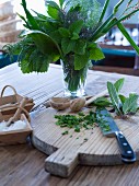 Various herbs in glass of water next to chopping board