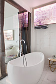 Free-standing bathtub with floor-mounted tap fittings in front of glass wall in ensuite bathroom