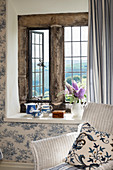 Cushion on wicker armchair in front of rustic stone window and toile de jouy wallpaper