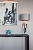 Silver lamp and ornament on console table below modern artwork on wall