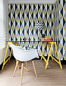 Table with glass top and shell chair in front of wall with geometric wallpaper