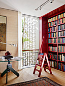 Red bookcase with matching ladder, seagrass wallpaper and round table in the library with oak parquet