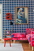 Pink, patterned sofa below painting on wall with blue, patterned wallpaper
