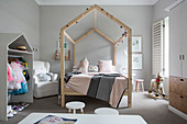 Four-poster bed with house-shaped wooden frame and open-fronted wardrobe in girl's bedroom with pale grey walls