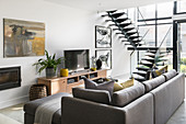 Grey sofa with scatter cushions on TV cabinet in open-plan interior with glass wall and staircase