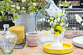 Narzissenstrauß in einer Krugvase, Ballonflaschen, Bindfaden, Pflanzentopf und Zinkeimer auf Gartentisch