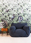 Quilted designer armchair and small retro table against wallpaper with Oriental design