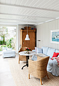 Sofa with pale blue loose cover, round table, wicker armchair and wooden cupboard in conservatory