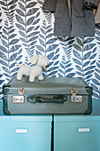 Pale blue storage boxes, suitcase and dog figurine in cupboard with patterned wallpaper on back wall