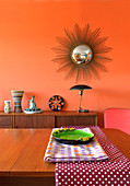 View across dining table to sideboard below sunburst mirror on orange wall