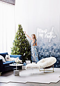 Blonde Frau dekoriert Weihnachtsbaum in Zimmerecke mit gemütlichen Sitzmöbeln