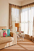 Light upholstered sofa with cushions and side table with lamp next to window