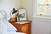 Tischlampe, Blumenvasen und gerahmtes Bild auf rustikalem Holz-Nachtkästchen, neben Bett